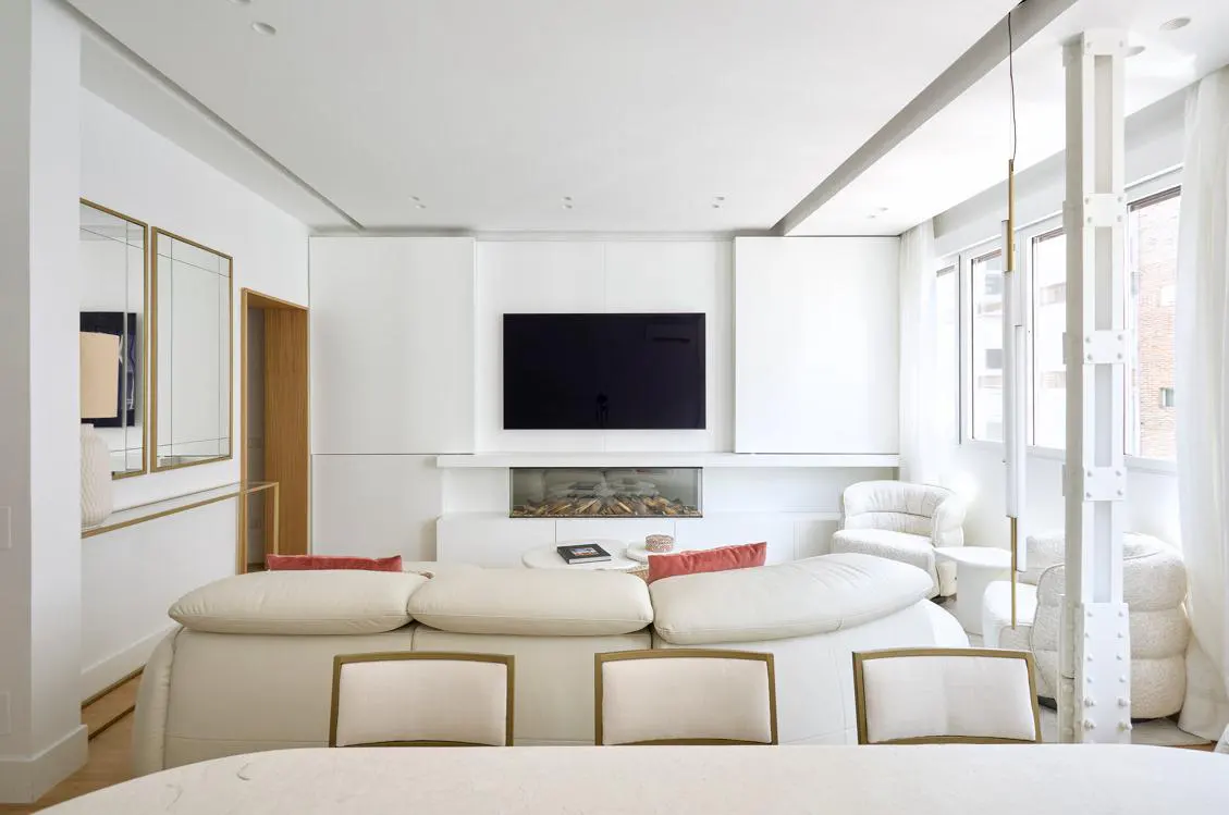 Bright, modern living room with white walls, furniture, and a large TV above a linear fireplace. Windows with sheer curtains let in natural light.