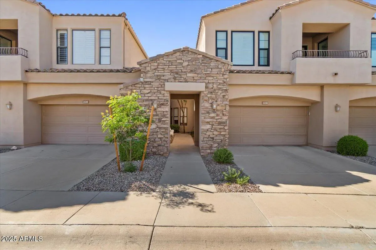 A tan townhouse with a stone entryway, a garage, and a small tree in front.