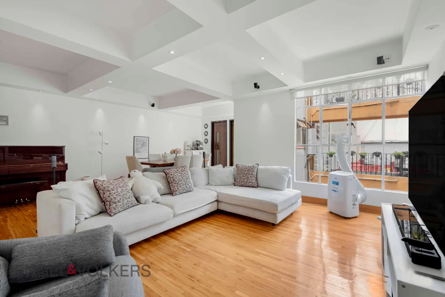 Bright living room with white walls, wood floors, and a large window. A white sectional sofa faces a TV. A piano and dining table are in the background.