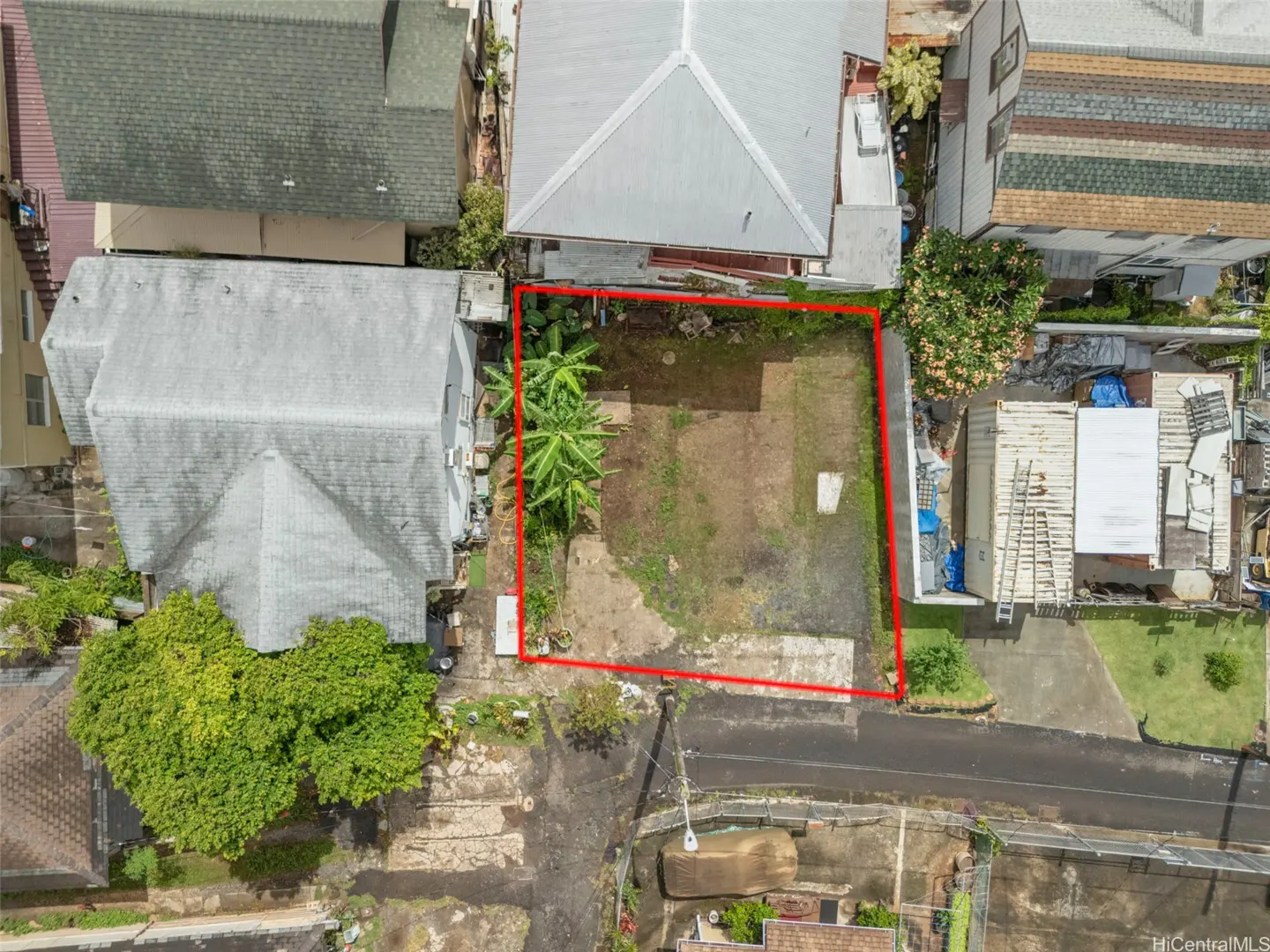 Aerial view of a vacant lot outlined in red, surrounded by houses and a street. The lot has some grass, trees, and a concrete slab.