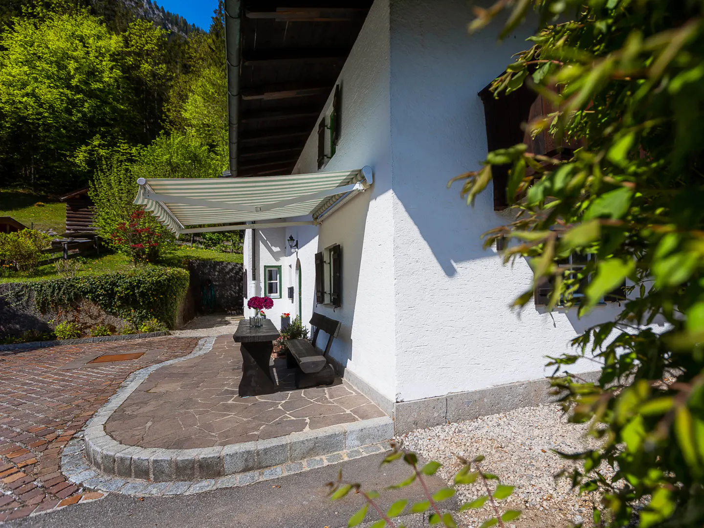 Exterior view of a white house with dark shutters, a striped awning, and a stone patio with a table and bench. Lush greenery surrounds the house.