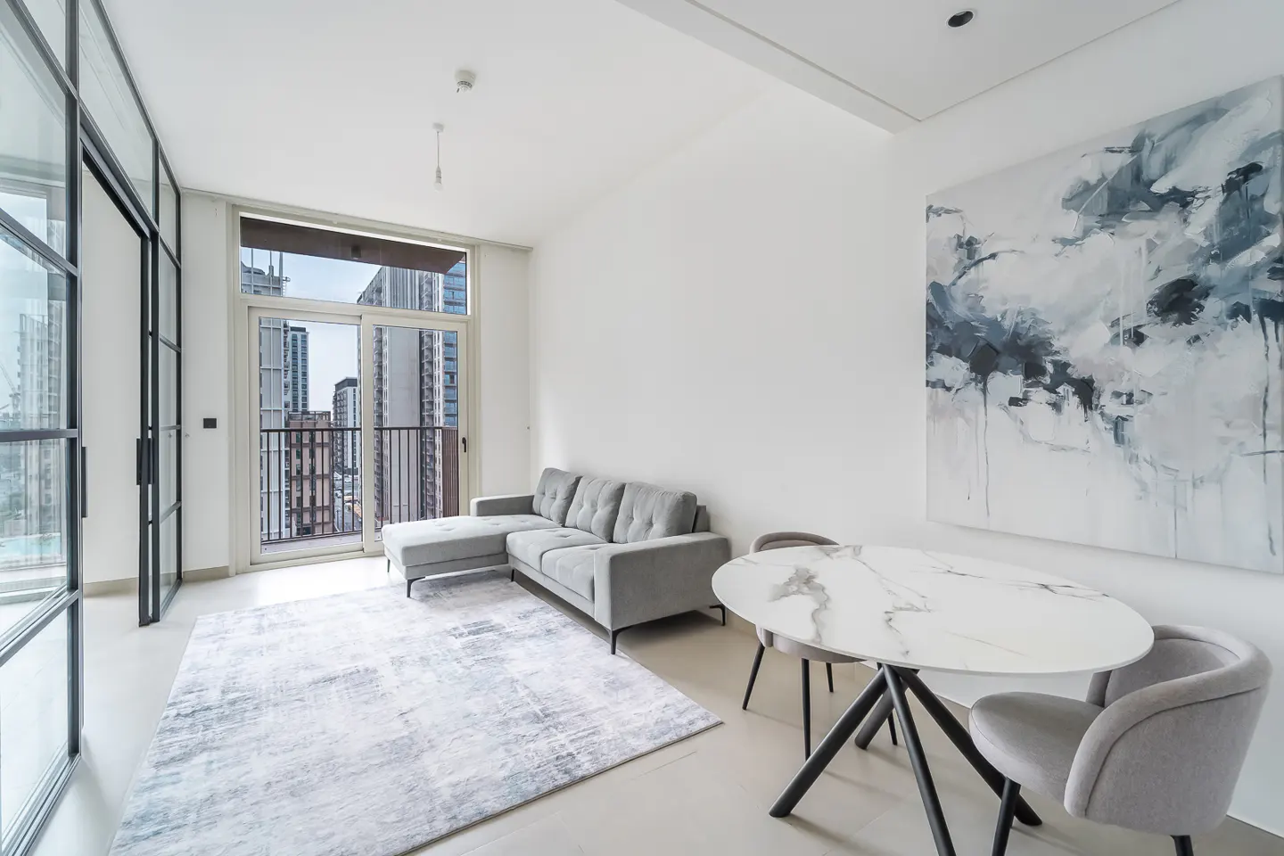 Bright living room with a gray sofa, marble table, and city view through black-framed windows. An abstract painting hangs on the white wall.