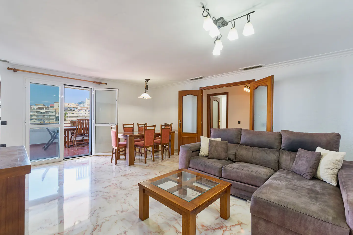 Bright living room with marble floors, brown sectional sofa, glass-topped coffee table, and dining table with red chairs. Balcony with city view.