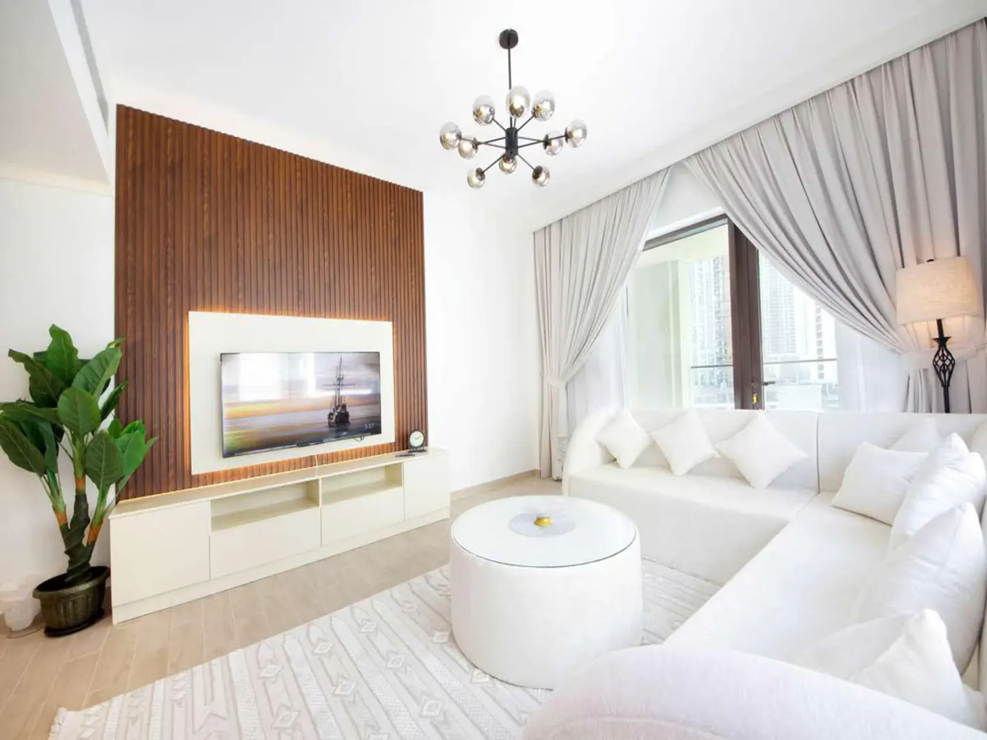 Bright living room with white sofa, round table, and TV on a wood-paneled wall. A modern chandelier hangs from the ceiling. Curtains frame a window.