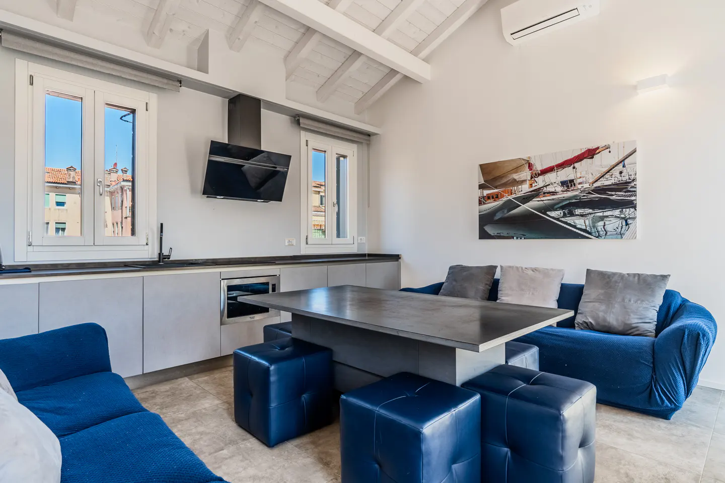 Modern kitchen and living area with blue sofas, a central dining table, grey cabinets, and a sailboat wall art piece.