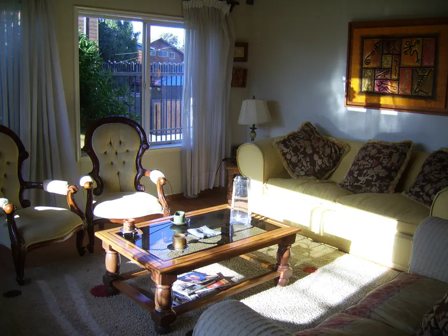 Sunlit living room with two armchairs, a wooden coffee table, and a yellow sofa with patterned pillows. A window shows a view of a house and trees.
