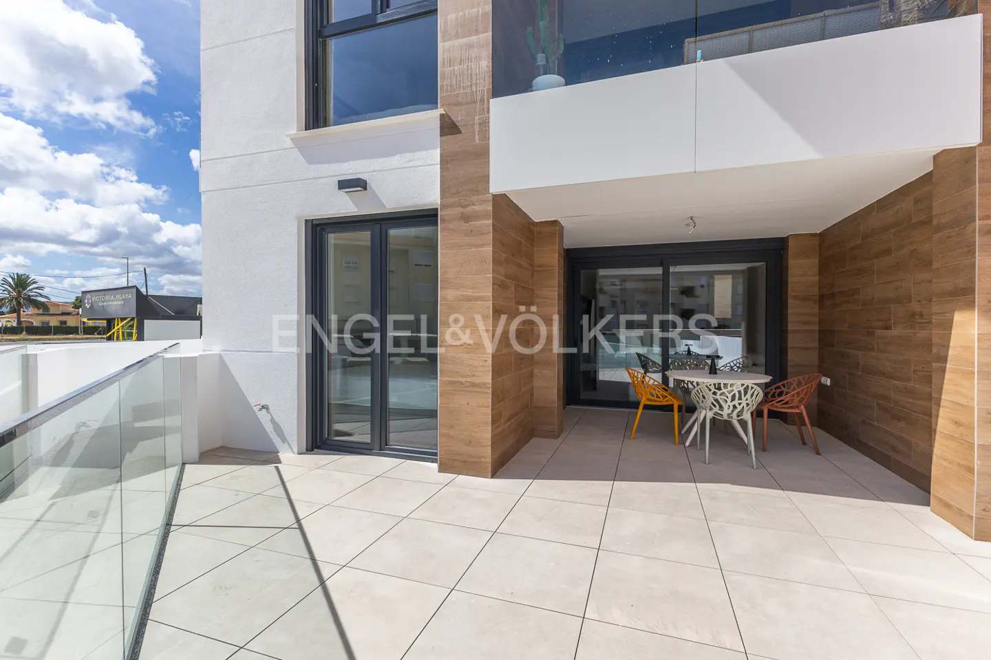 Exterior view of a modern apartment balcony with a table and chairs, tiled floor, and glass railing. The building is white with wood-look accents.