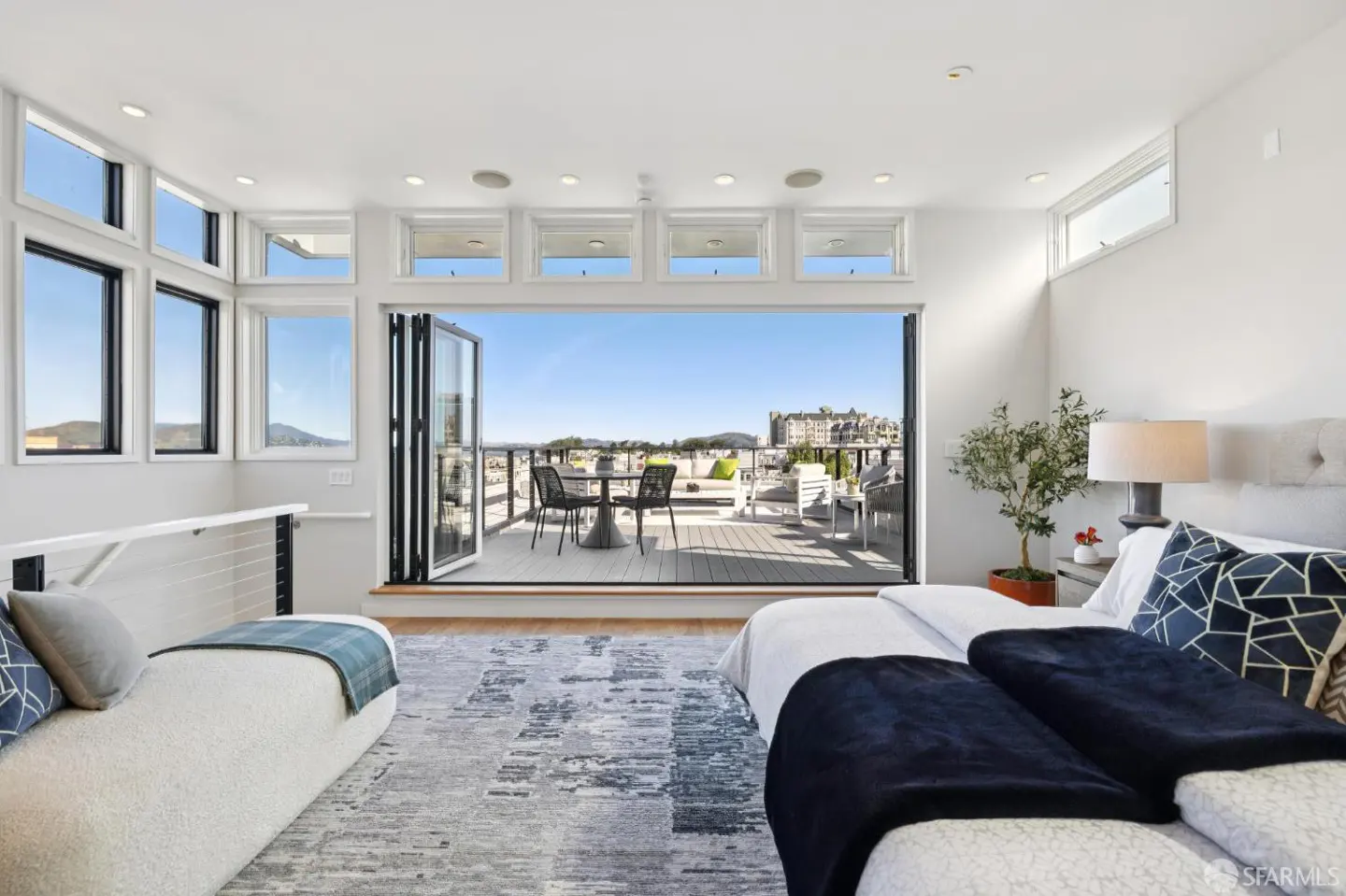 Bright bedroom with a large folding door opening to a deck with city views. White walls, bed with blue accents, and a chaise lounge.
