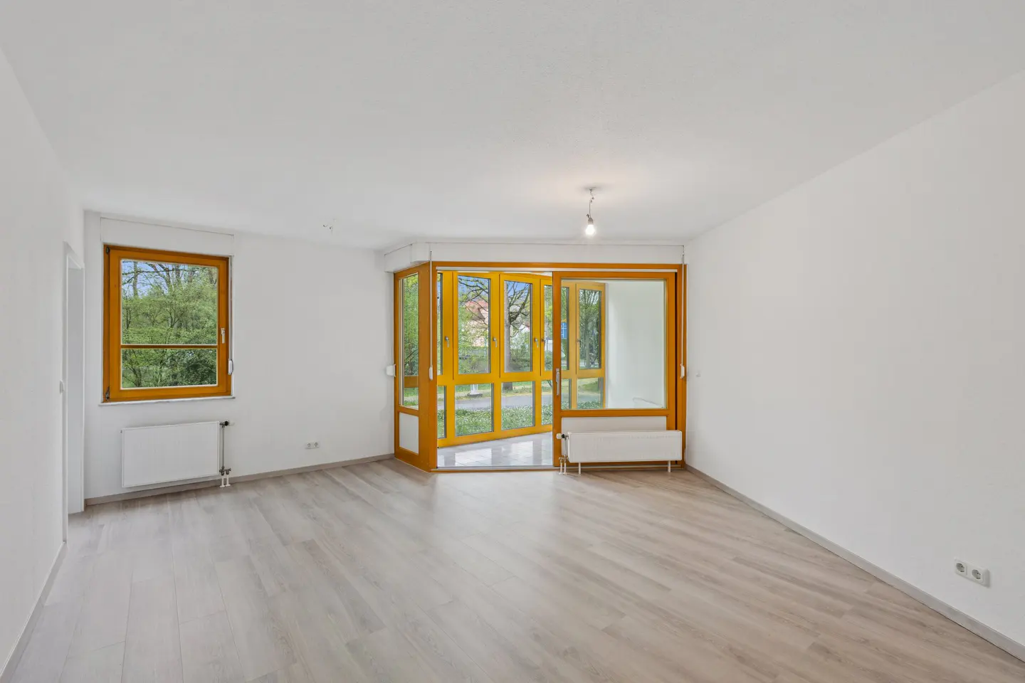 Bright, empty room with white walls, light wood floors, and a window and sliding glass doors with wood frames.