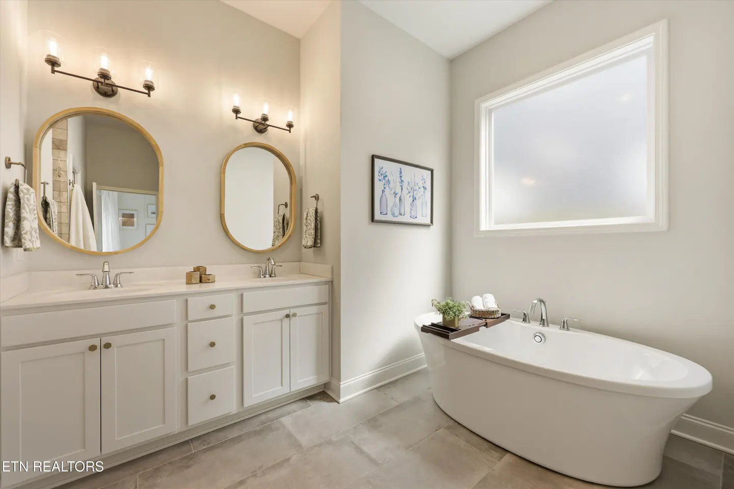 Bright bathroom with white cabinets, double sinks, and oval mirrors. A white tub sits beneath a frosted window. Gray tile floor.