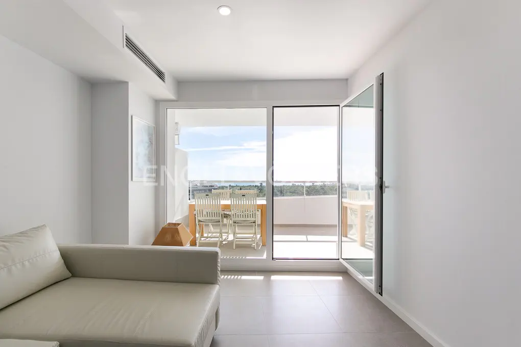 Bright, modern living room with white walls, a light sofa, and an open glass door to a balcony with outdoor seating and a view.
