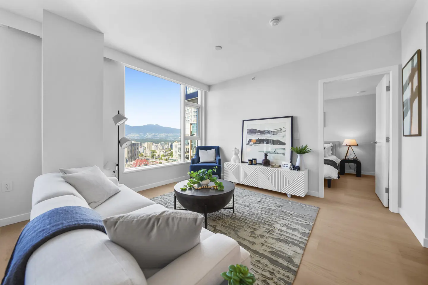 Bright, modern living room with a white sofa, blue chair, and city view. A round coffee table sits on a gray rug. Bedroom visible through doorway.