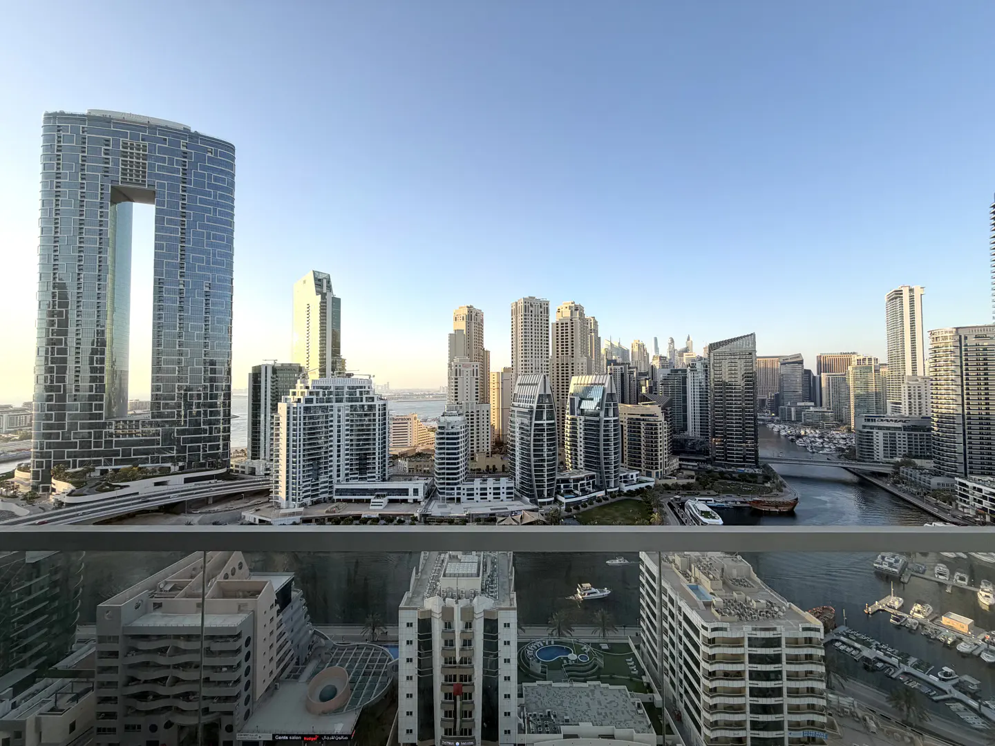 High-angle view of the Dubai Marina skyline at sunset, featuring modern skyscrapers, a canal with boats, and a clear sky.