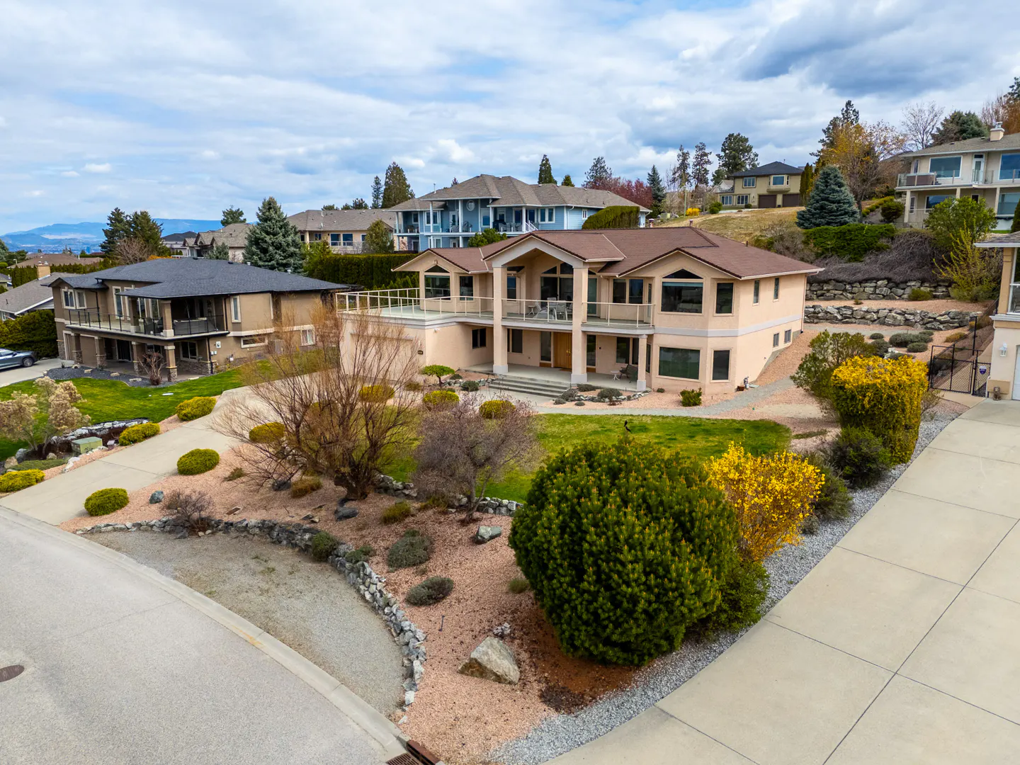 Aerial view of a two-story beige house with a brown roof, a balcony, and a landscaped yard on a cloudy day.