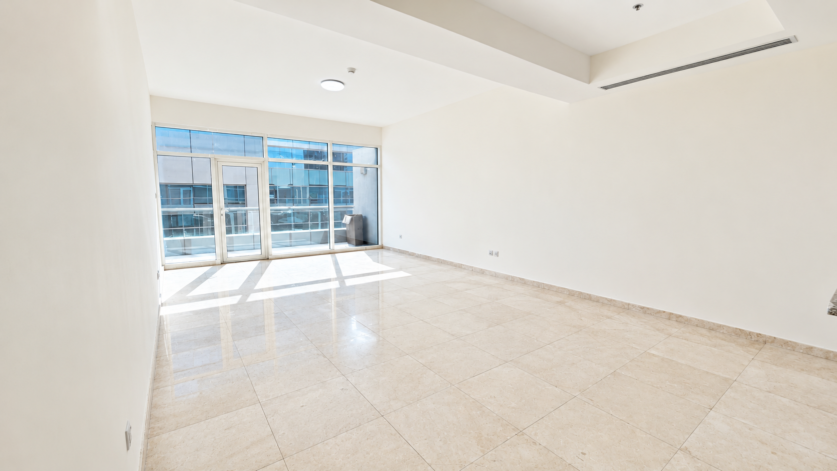 Empty, bright room with white walls, polished beige tile floors, and large glass doors leading to a balcony. [AI-generated text]