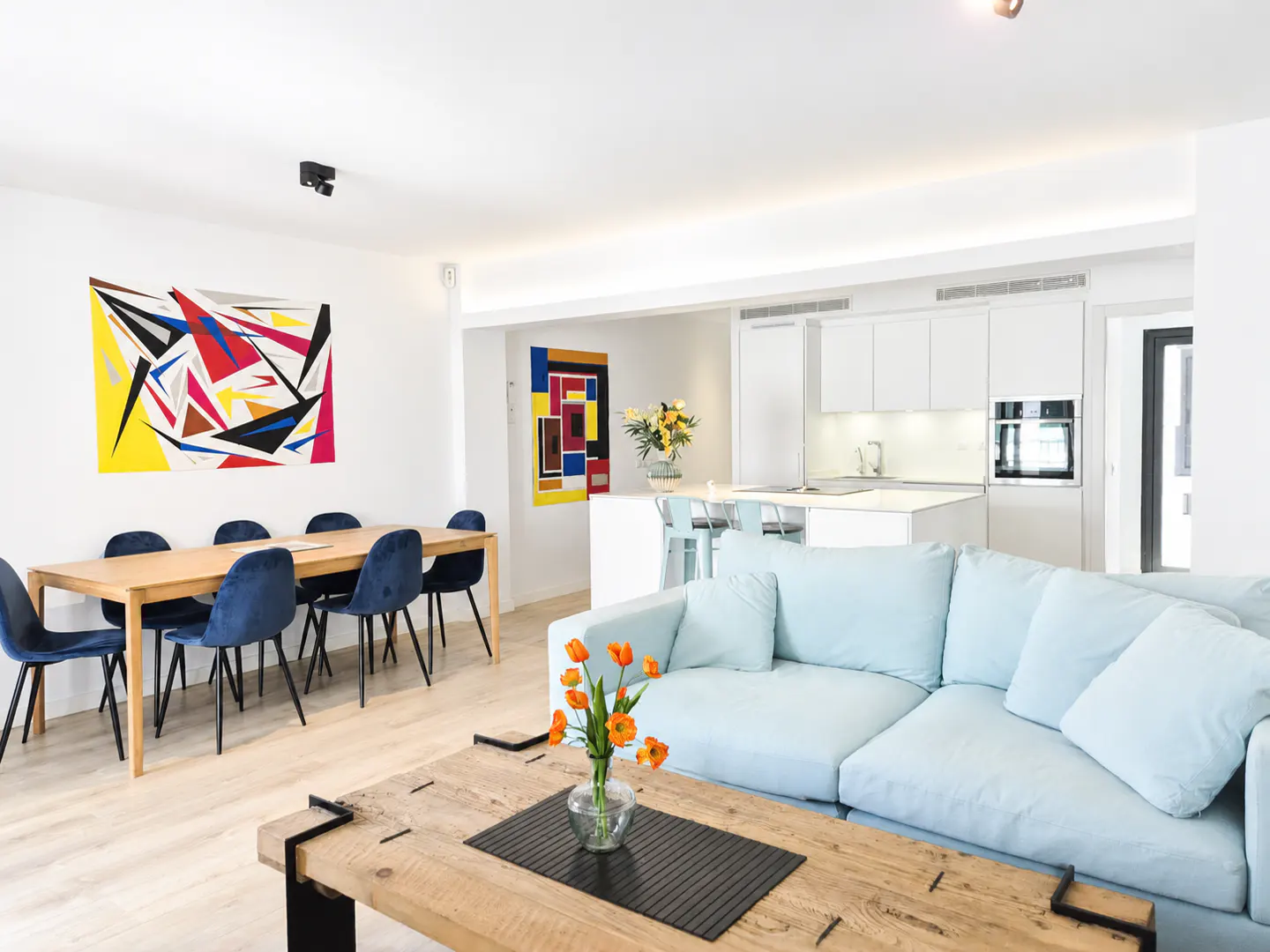 Bright, open-concept living space with a light blue sofa, wooden coffee table, and dining table with blue chairs. Modern art adorns the white walls.