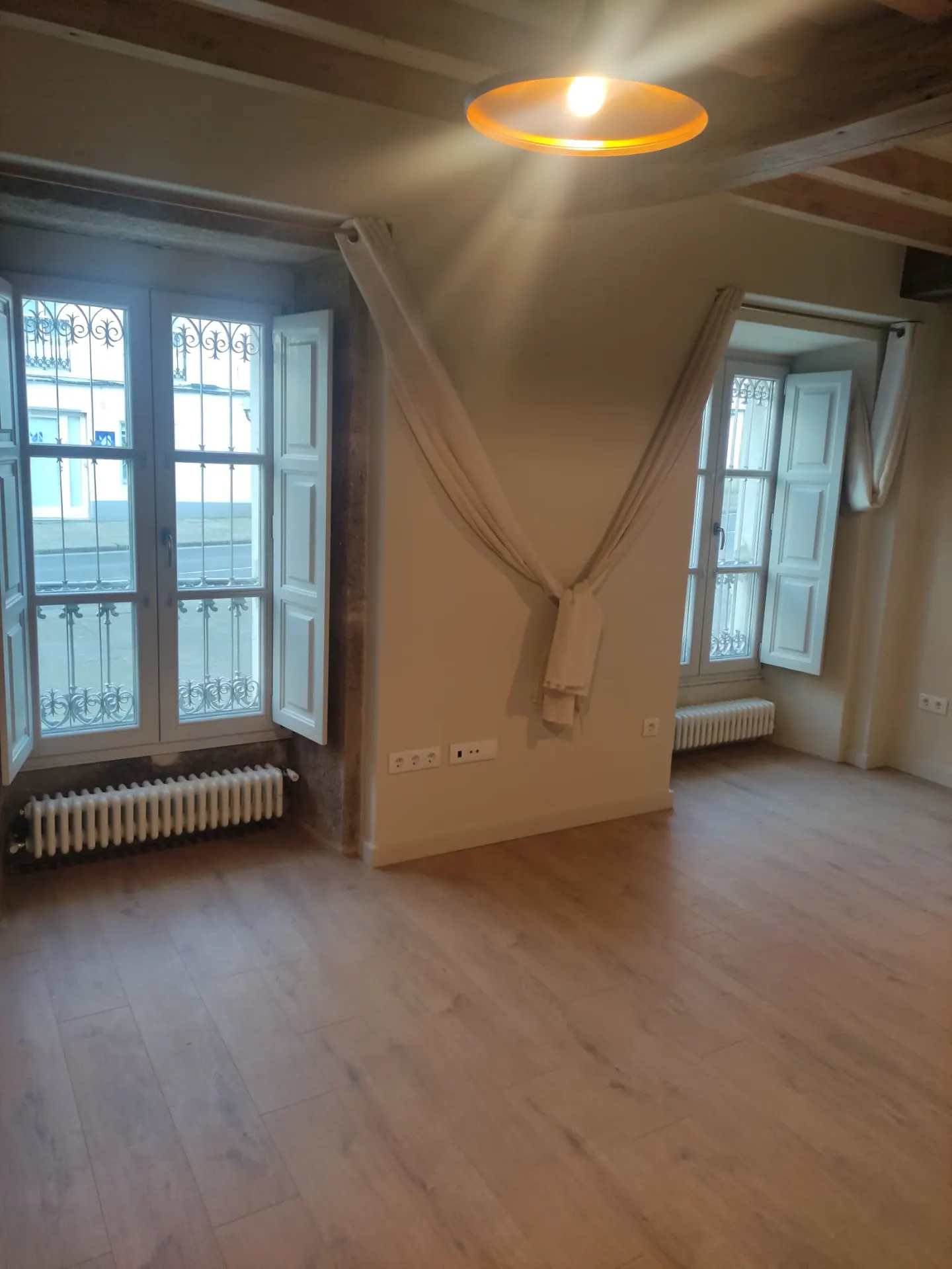 Empty room with light wood floors, beige walls, and two windows with white shutters. A beige curtain is draped on the wall. A round light fixture is on the ceiling.