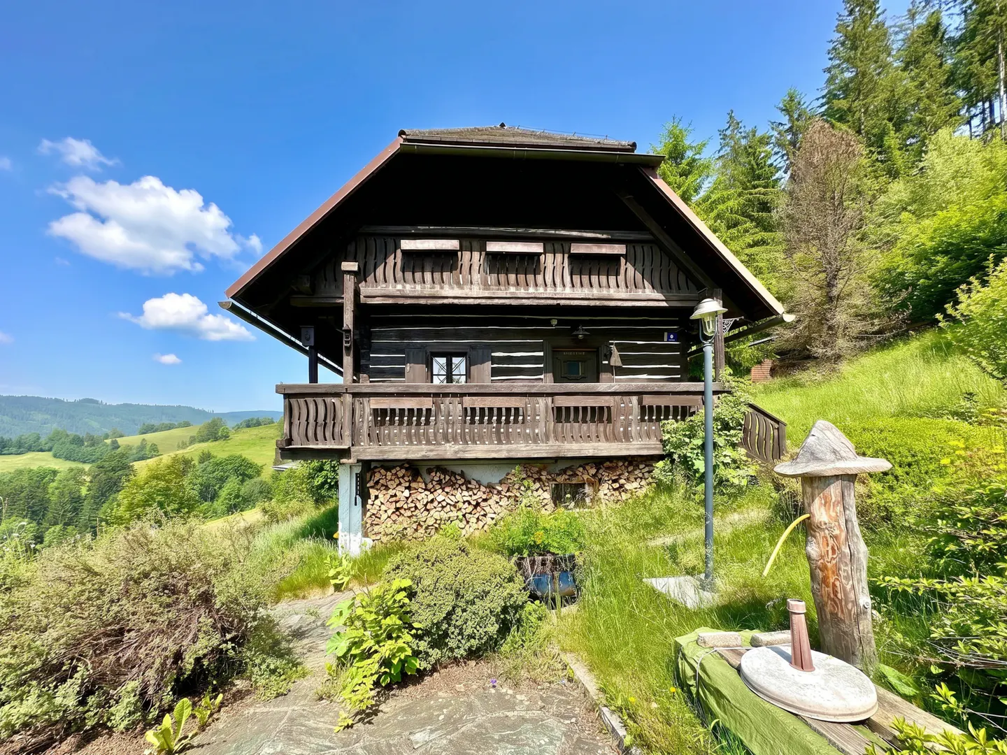 Rustic log cabin with a balcony, nestled on a green hillside under a blue sky. Firewood is stacked beneath the house.