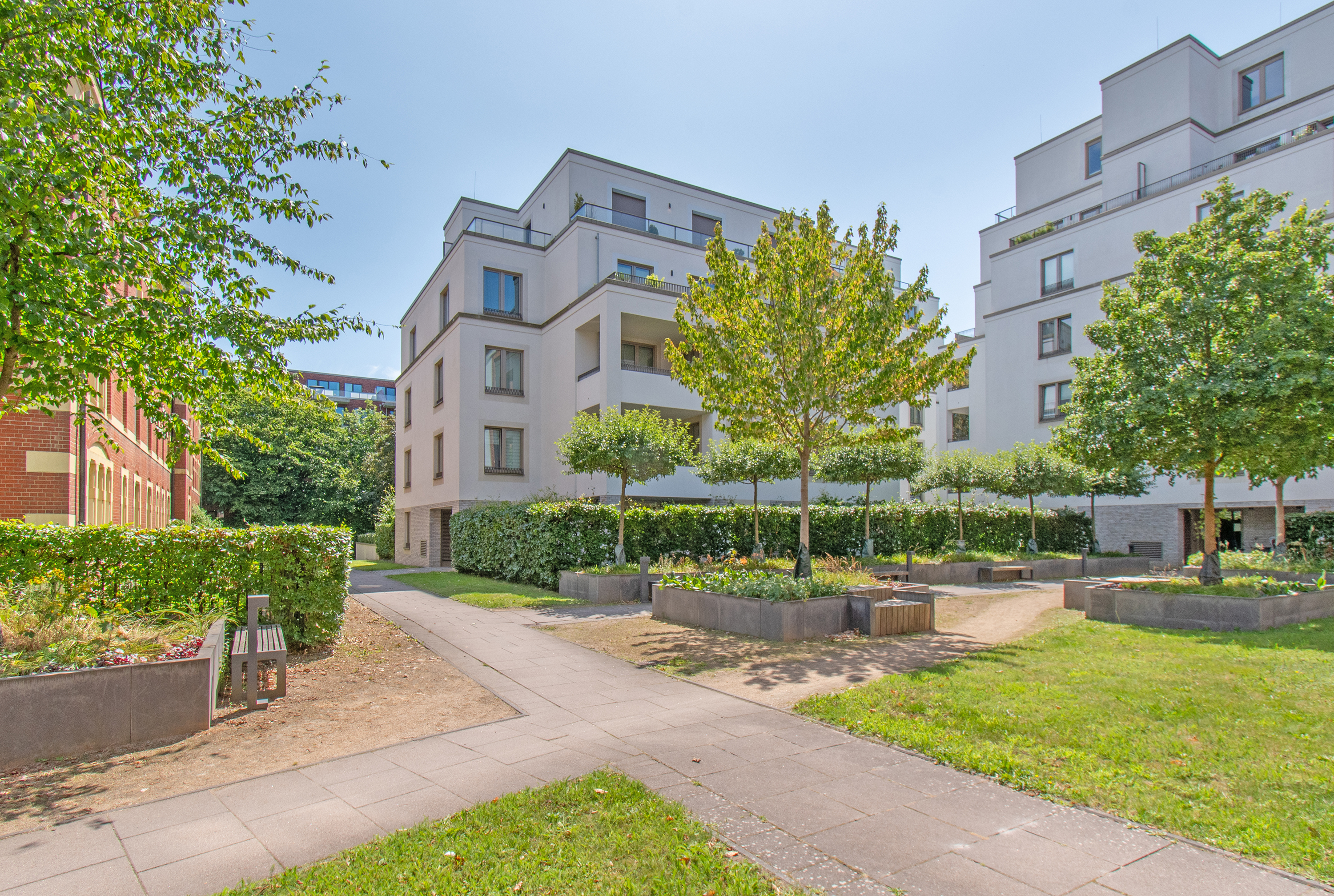 Modern white apartment buildings surrounded by a landscaped courtyard with paved paths, green trees, and manicured hedges. [AI-generated text]