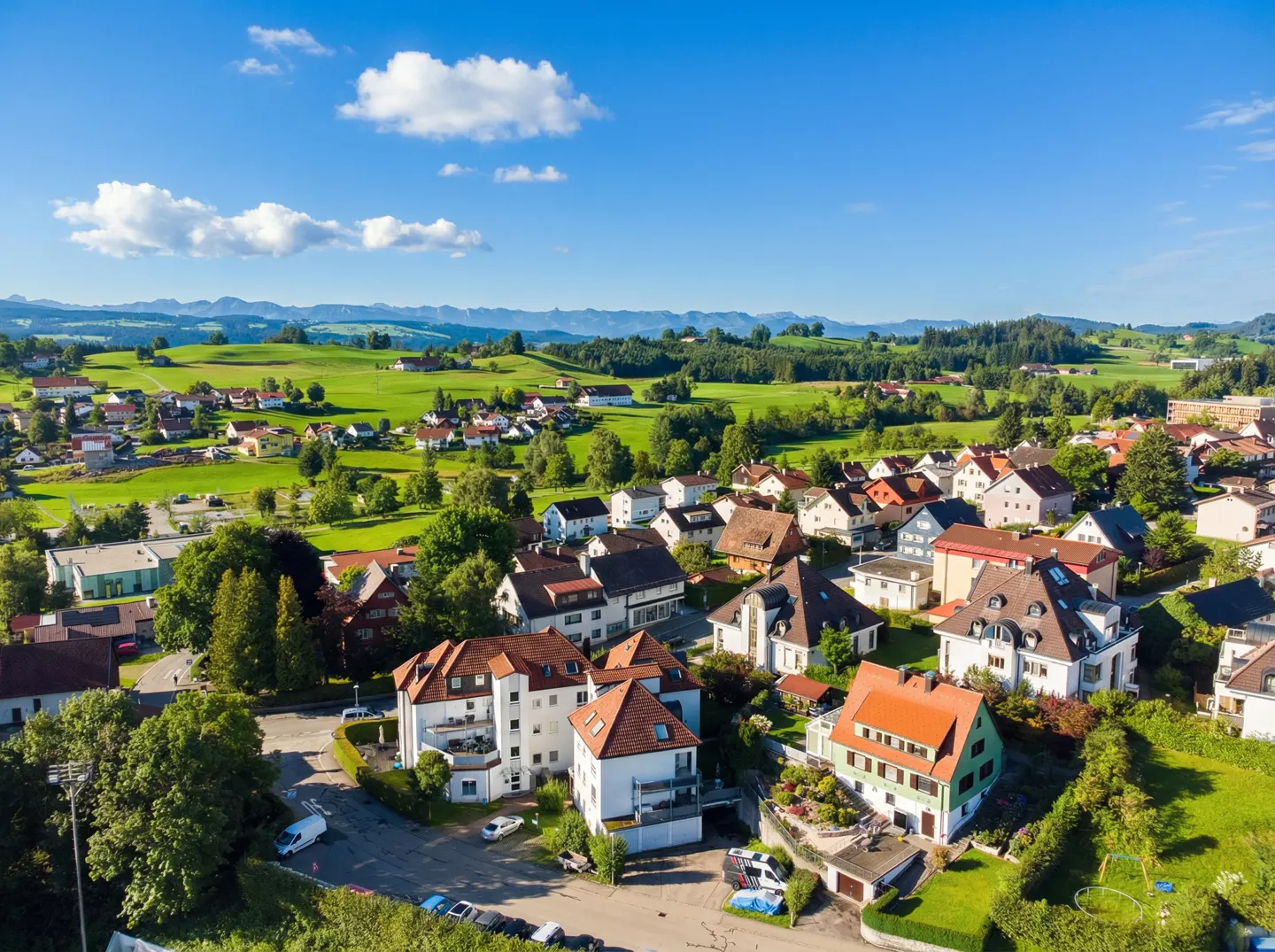 Aerial view of a European village with red-roofed houses, green fields, and distant mountains under a blue sky with white clouds.