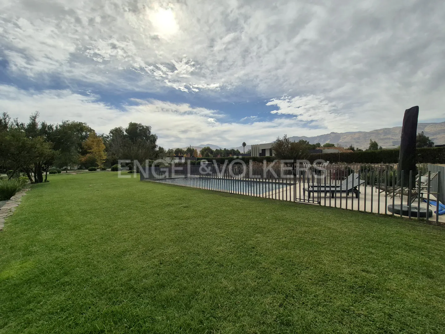 A wide shot of a green lawn, pool with fence, lounge chairs, and mountains in the background.