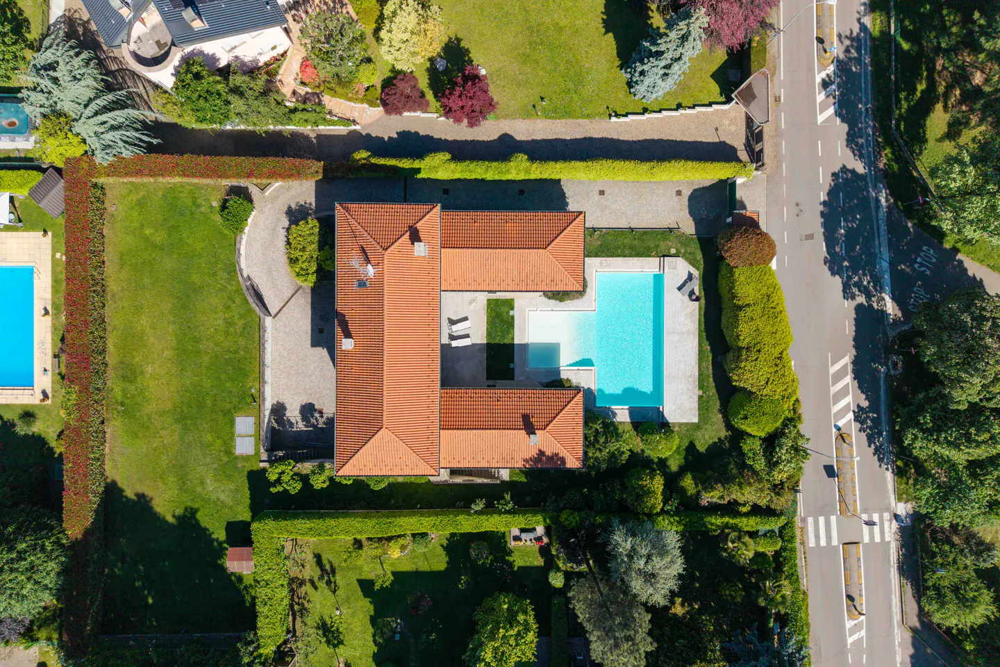 Aerial view of a terracotta-roofed house with a pool, surrounded by green lawns and hedges. A road runs along one side.