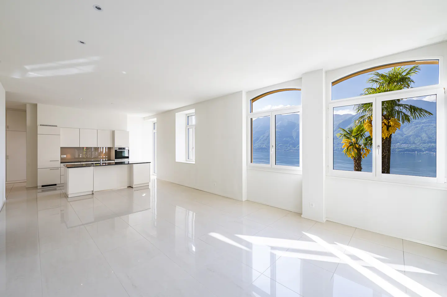 Bright, modern apartment interior with white walls, tile floors, and a kitchen. Large windows offer a view of palm trees, mountains, and a lake.