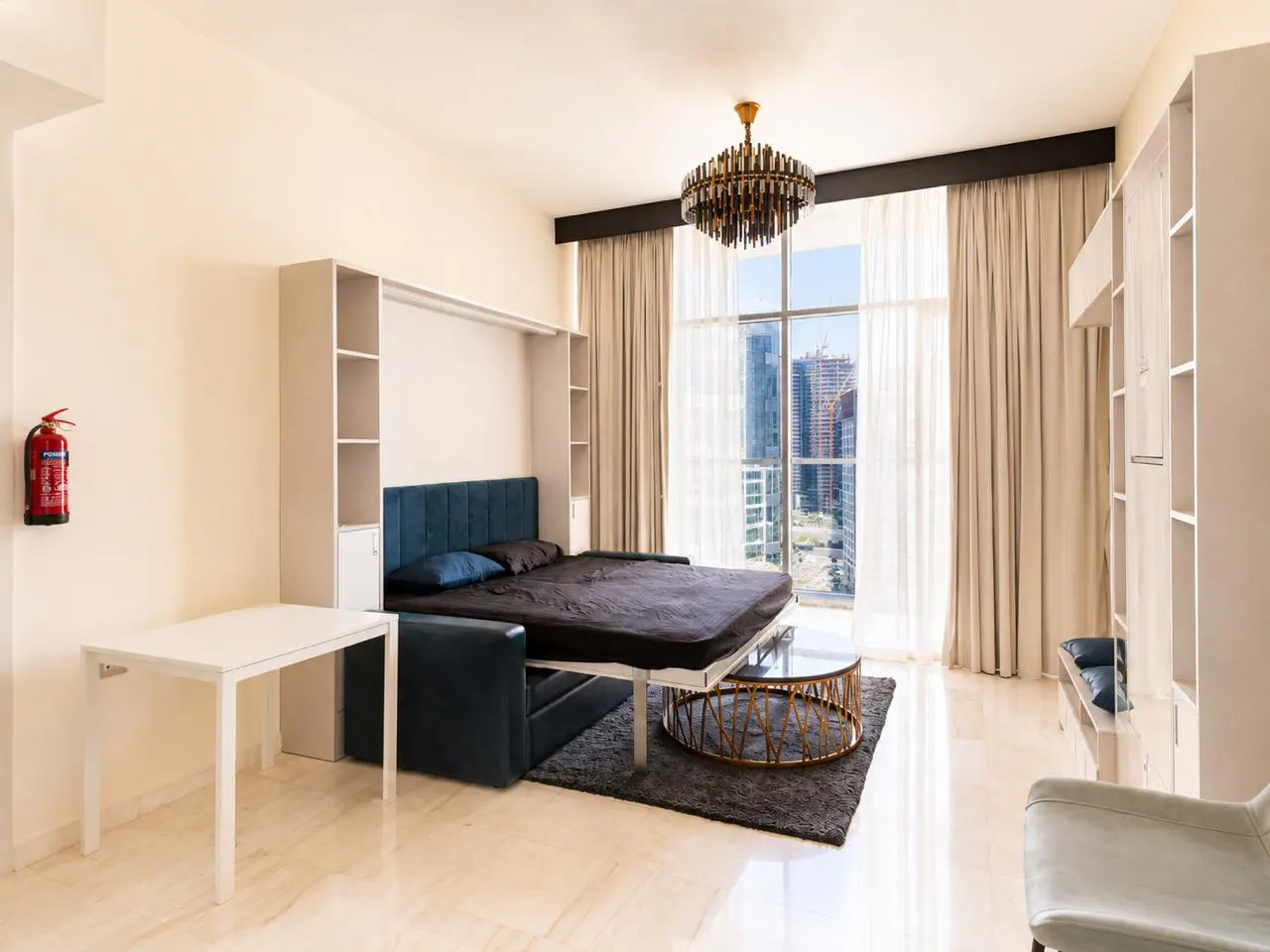 Bright studio apartment with a blue sofa bed, white shelving, and a city view from a large window. A round coffee table sits on a gray rug.