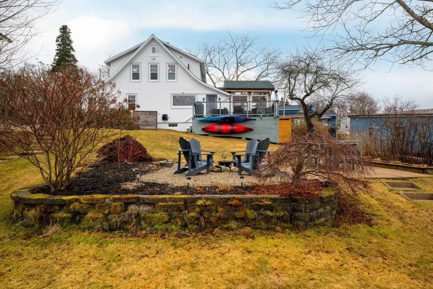 Backyard view of a white house with a deck, kayaks, and a stone patio with chairs.