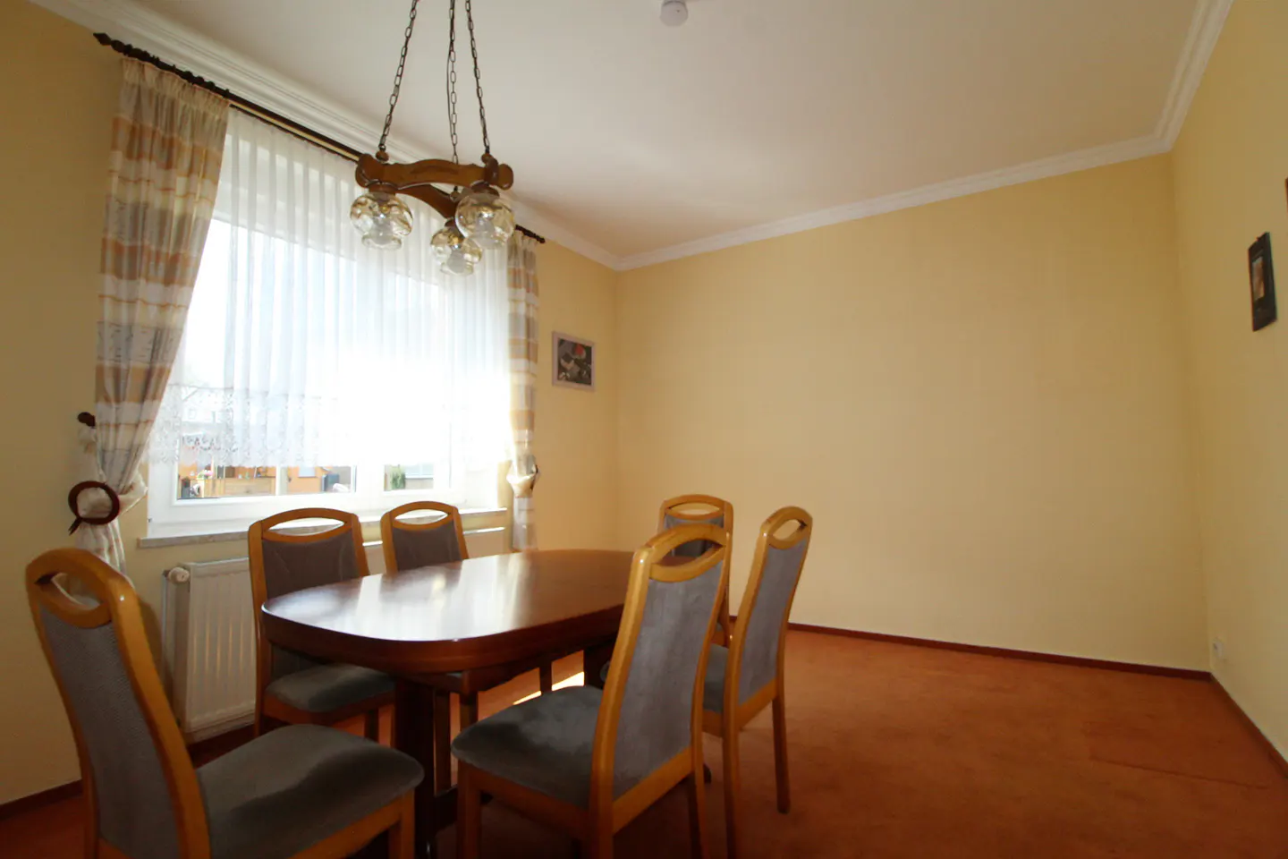 Dining room with a wooden table, six chairs, and a chandelier. The walls are yellow, and the floor is carpeted in brown. Curtains cover the window.
