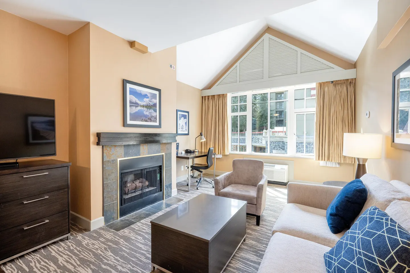 Living room with fireplace, TV, desk, and large window. Beige walls, neutral furniture, and blue accent pillows.