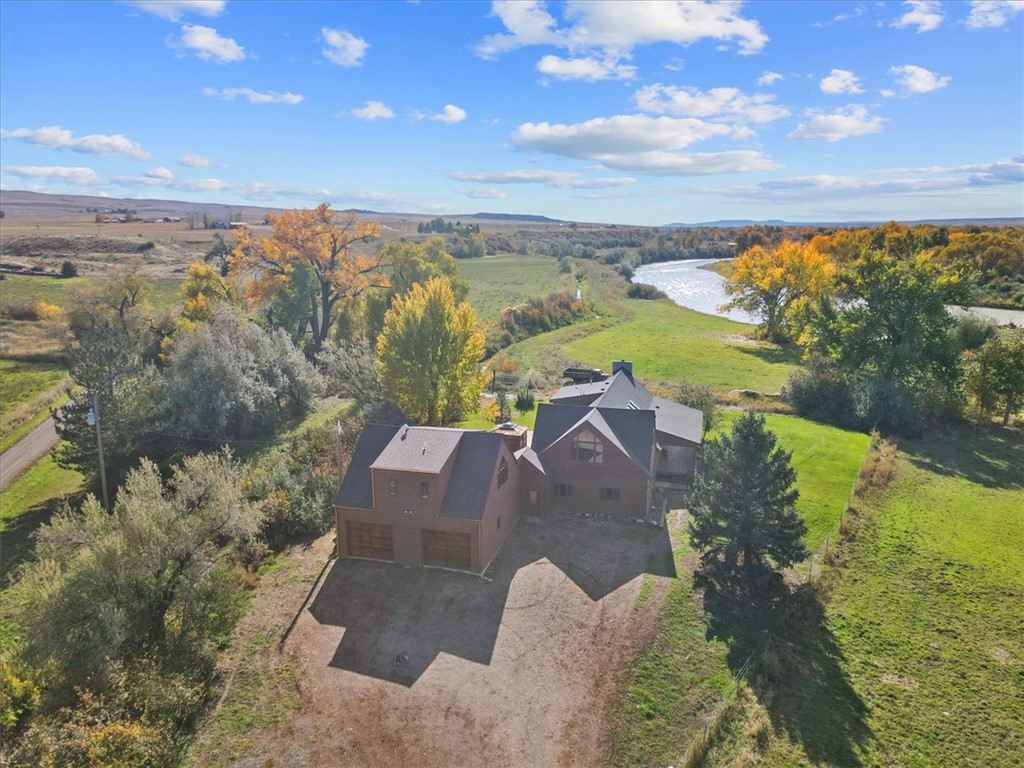 Aerial view of a brown house with a gray roof, surrounded by green grass, trees with yellow leaves, and a river. Blue sky with clouds.