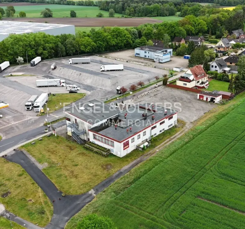 Aerial view of a white commercial building with a red roof, surrounded by parking lots, trucks, and green fields.