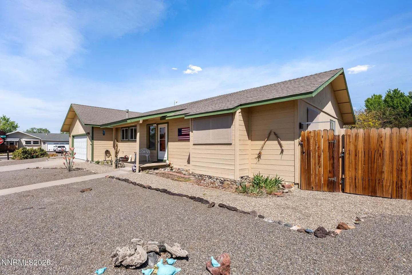 Tan, single-story home with a brown roof, green trim, and a wooden fence. An American flag hangs near the front door. The yard is covered in gravel.