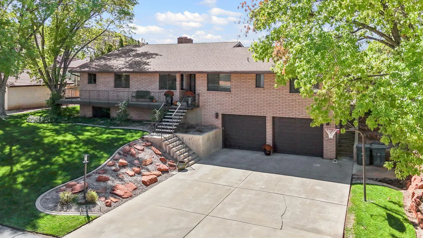 Two-story brick house with a brown roof, two-car garage, and a basketball hoop in the driveway. Green trees surround the house.