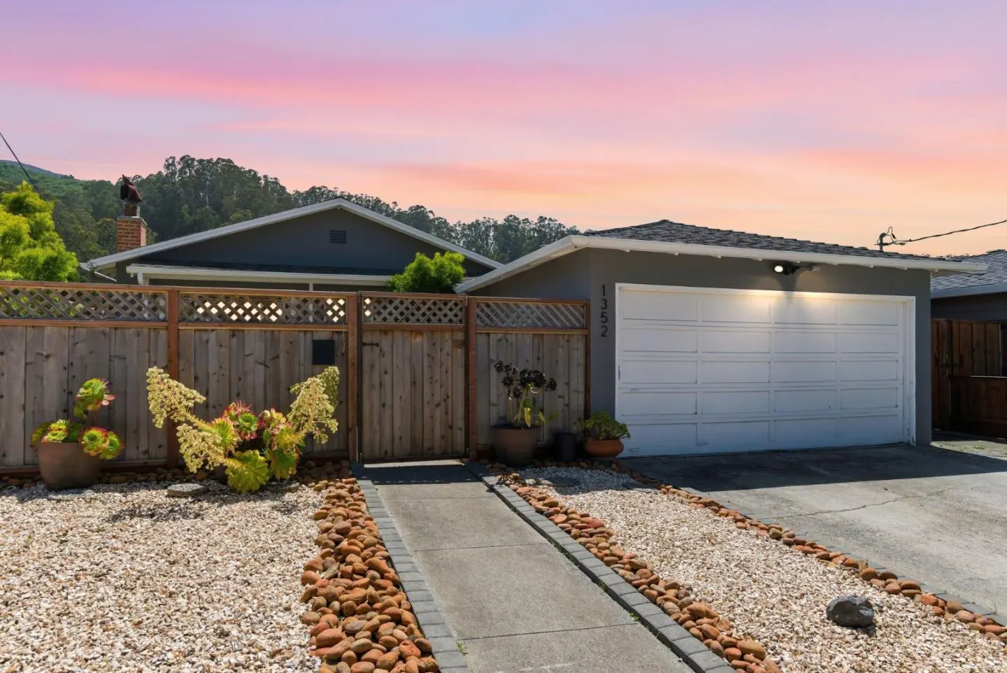 Exterior of a gray house with a white garage door, wooden fence, and gravel yard at sunset.