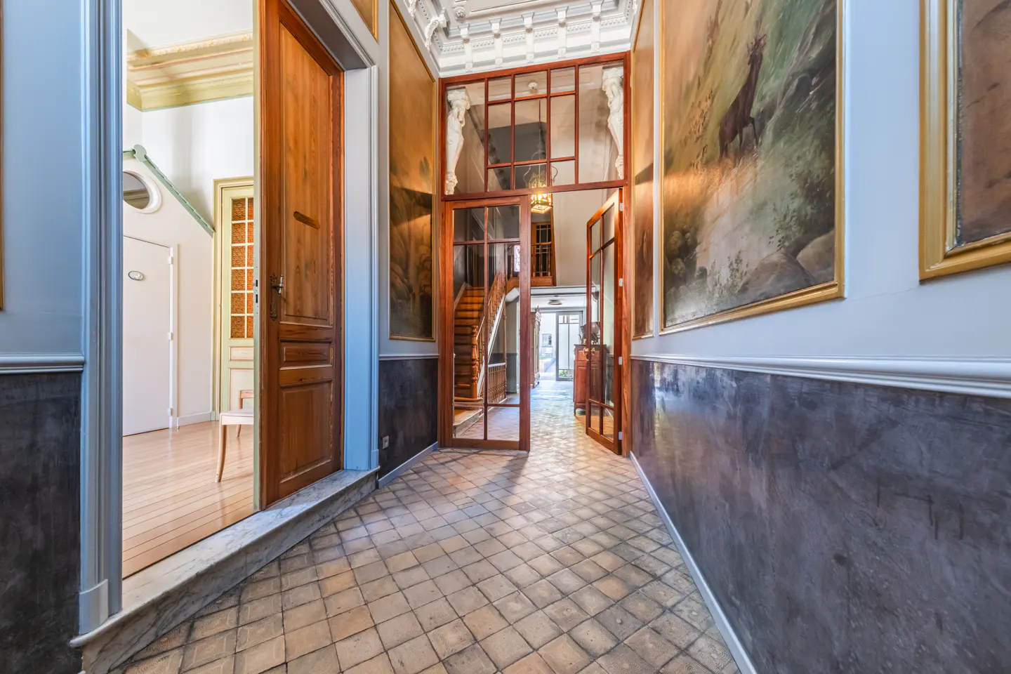 Hallway with tan tile floor, wood doors, and gray walls. Paintings hang on the walls. A glass door leads to a staircase.