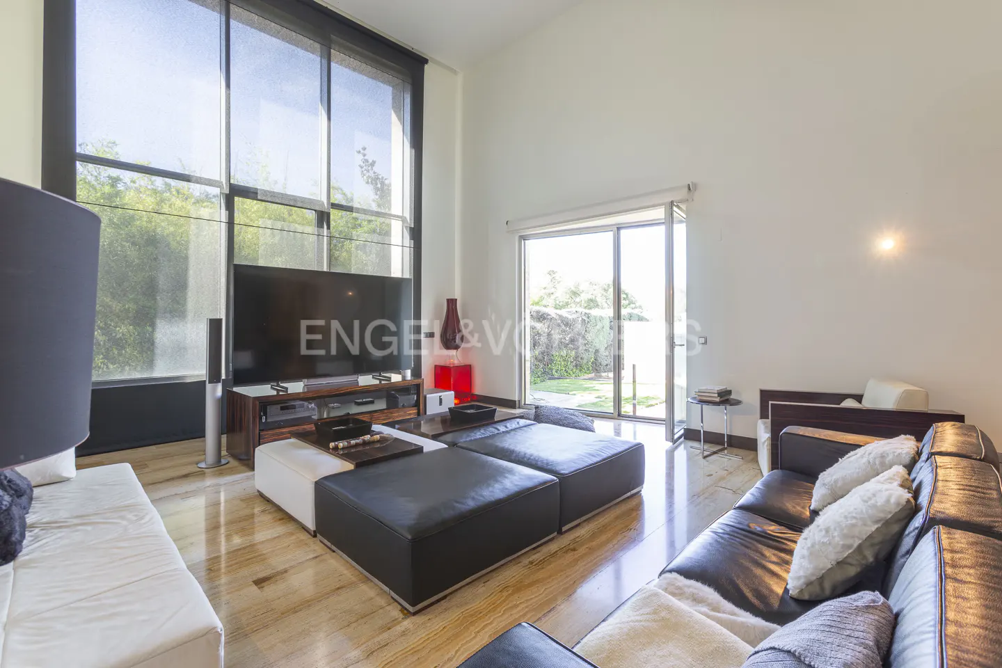 Bright living room with black leather sofa, white cushions, and black and white ottomans. Large windows overlook a green yard.