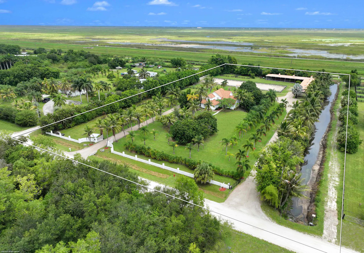 Aerial view of a large property with a white fence, palm trees, a house with a red tile roof, and a horse stable. Green fields in the background.