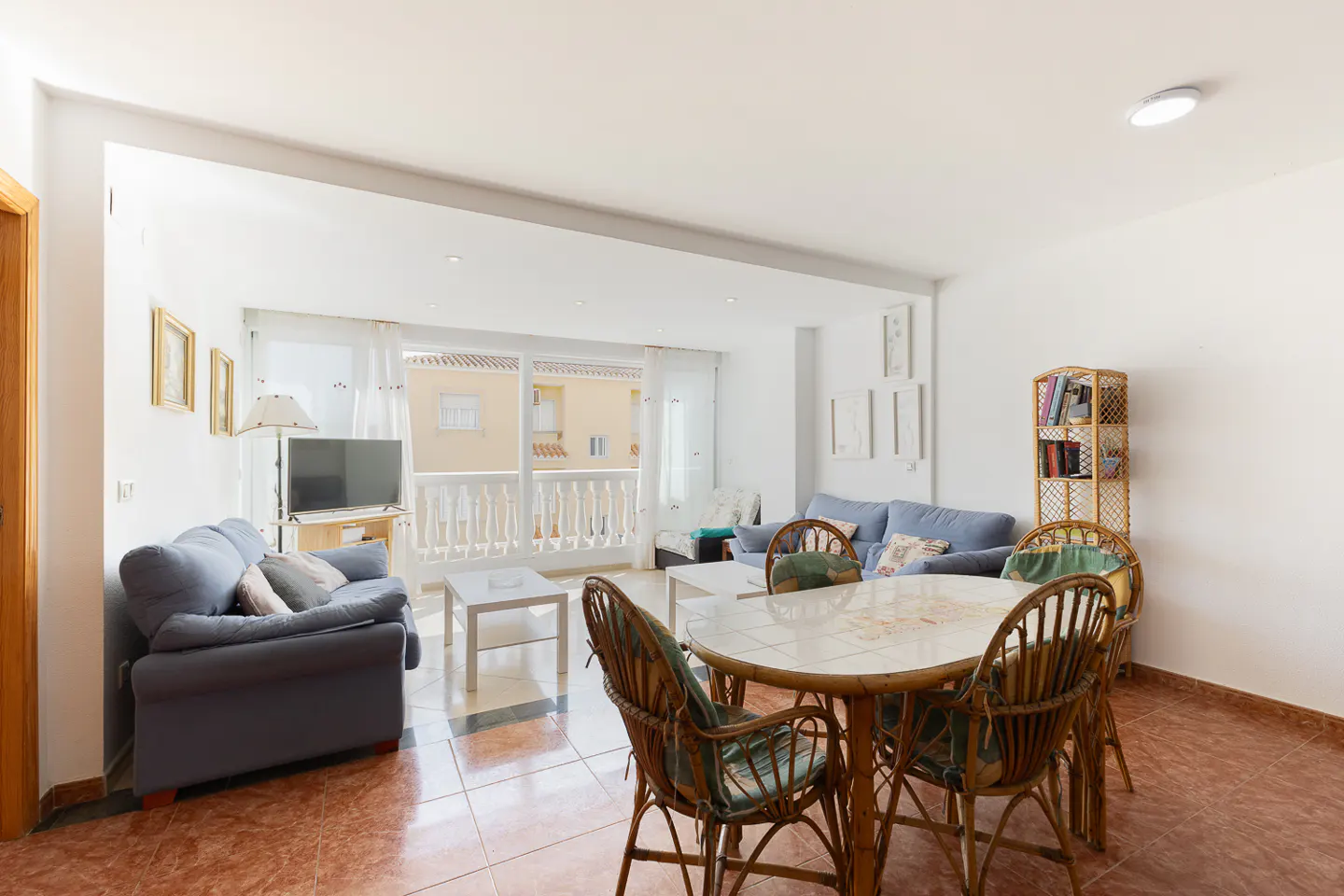 Bright living room with blue sofas, a round wicker dining table, and a balcony with white railings.
