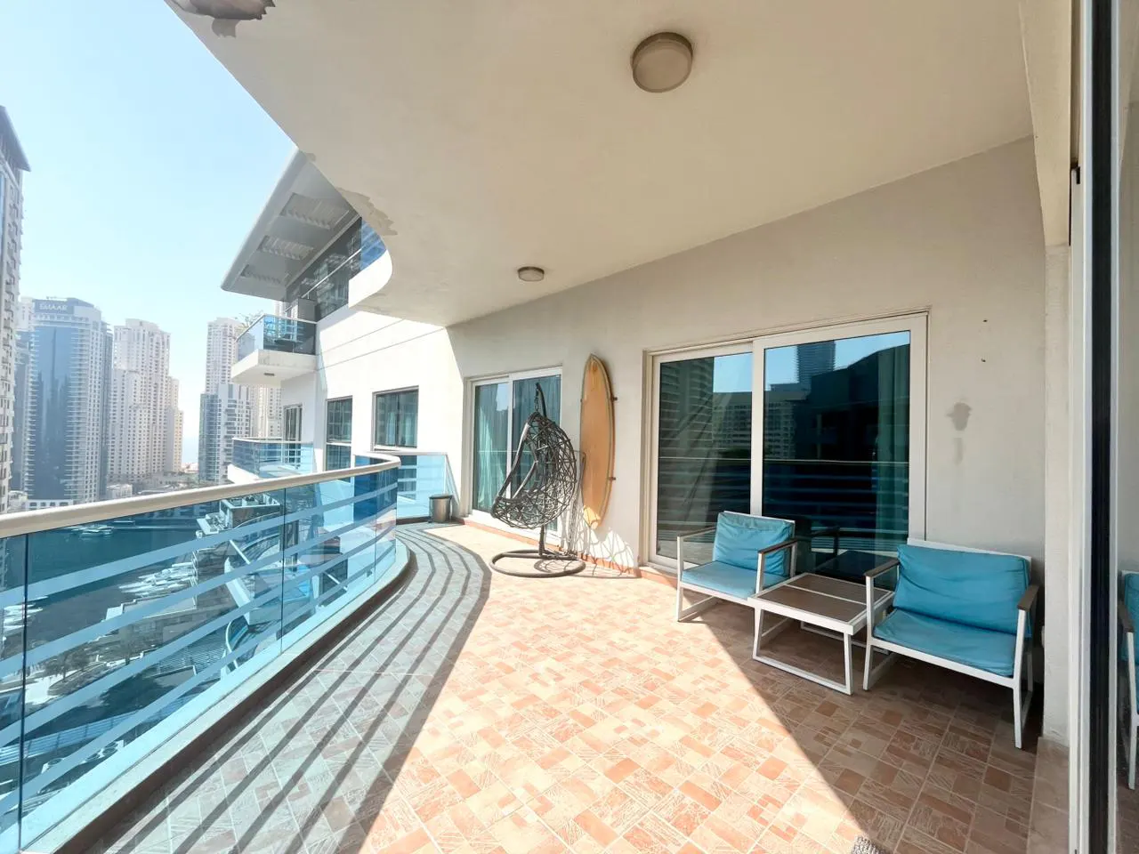 A condo balcony with blue glass railings overlooks a city skyline. The balcony has a hanging chair, surfboard, and blue cushioned seating.