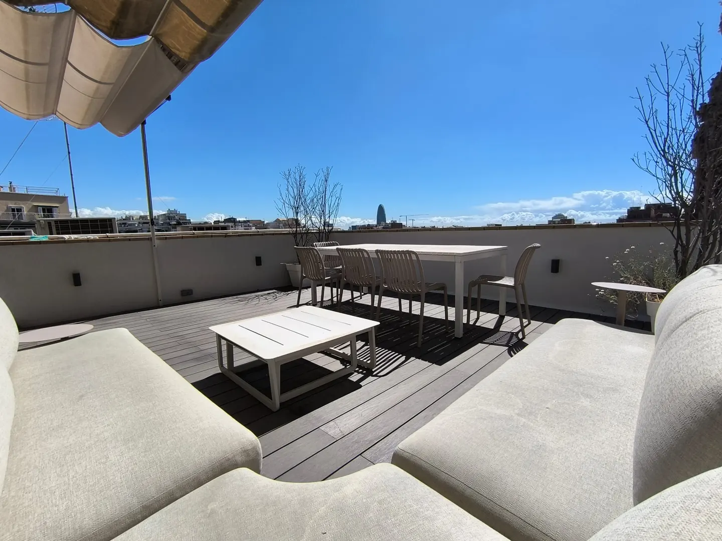 Rooftop patio with white furniture, including a table, chairs, and lounge seating. City skyline visible in the background under a bright blue sky.