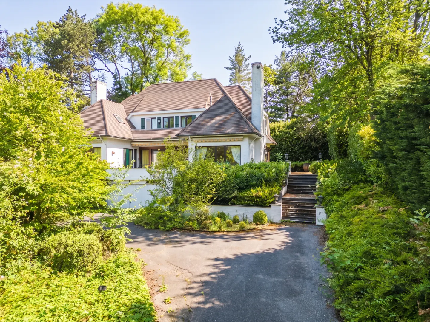 Exterior view of a white house with a brown roof, surrounded by green trees and bushes. A driveway leads to the house.