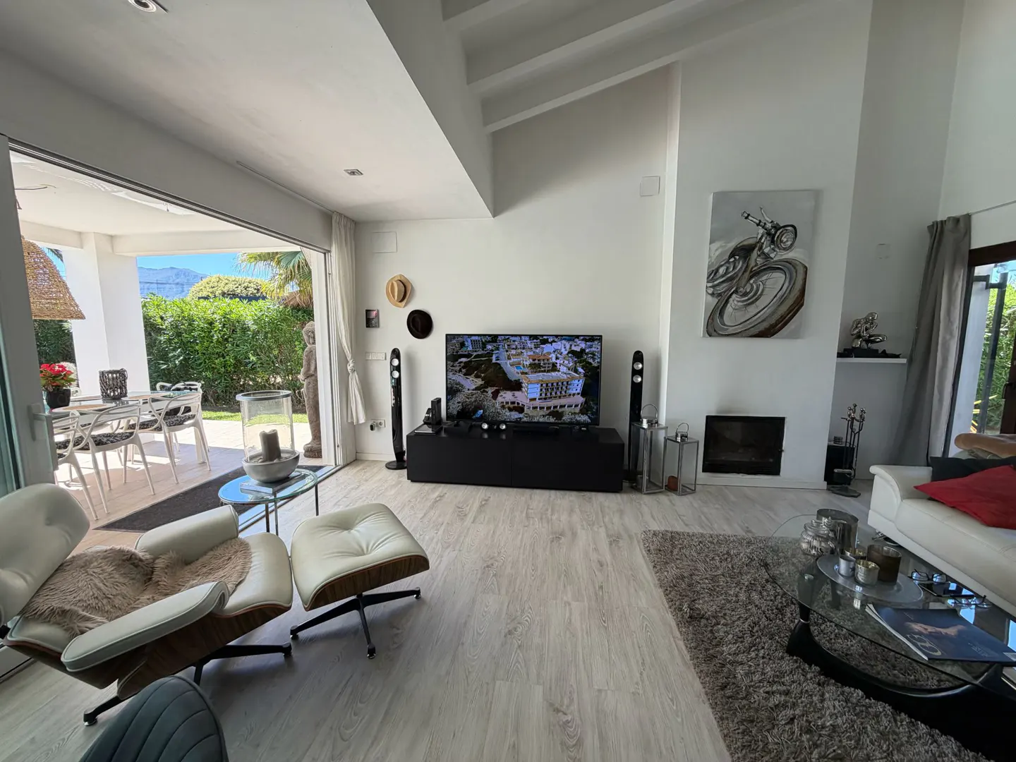 Bright living room with white walls, light wood floors, and open patio. A black TV console sits between speakers. A motorcycle painting hangs above a fireplace.