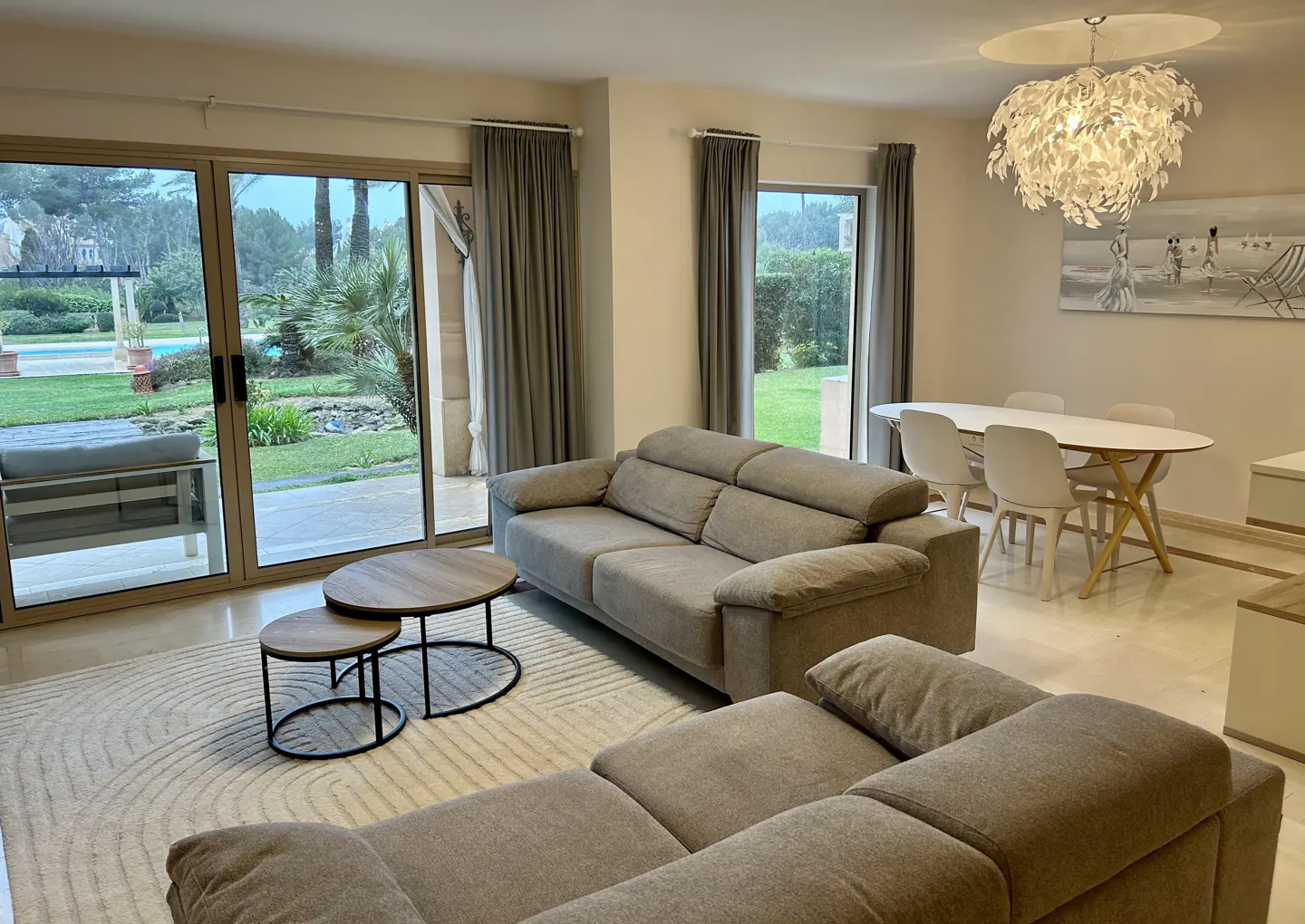 Living room with gray sofas, round tables, and a dining area with a white table and chairs. Sliding glass doors lead to a pool and garden.