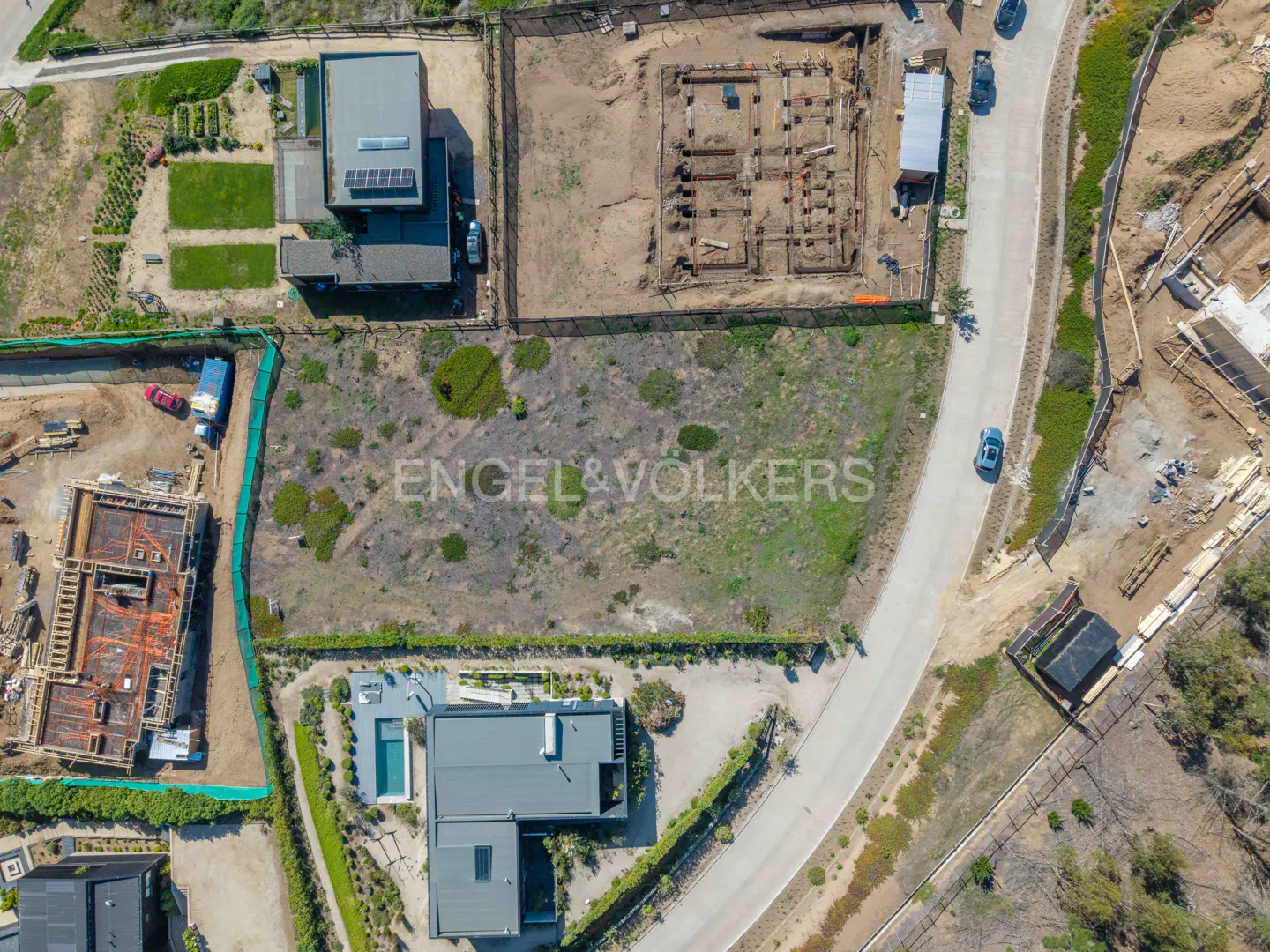 Aerial view of a vacant lot with Engel & Volkers logo, surrounded by modern homes, construction sites, and a winding road.