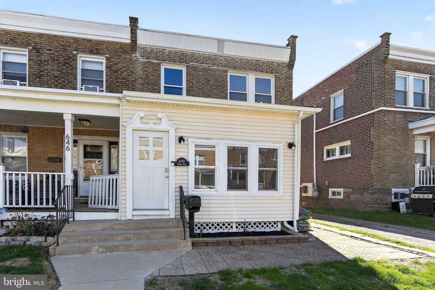 A two-story row house with a cream-colored facade and a white front door. A small porch with white railings is on the left. A black mailbox is on the right.