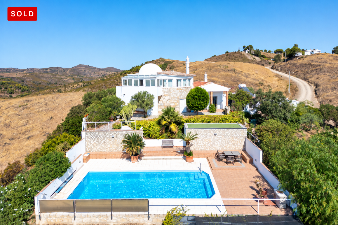 Aerial view of a white villa with a pool, stone walls, and a patio, set in a dry, hilly landscape under a clear blue sky. [AI-generated text]