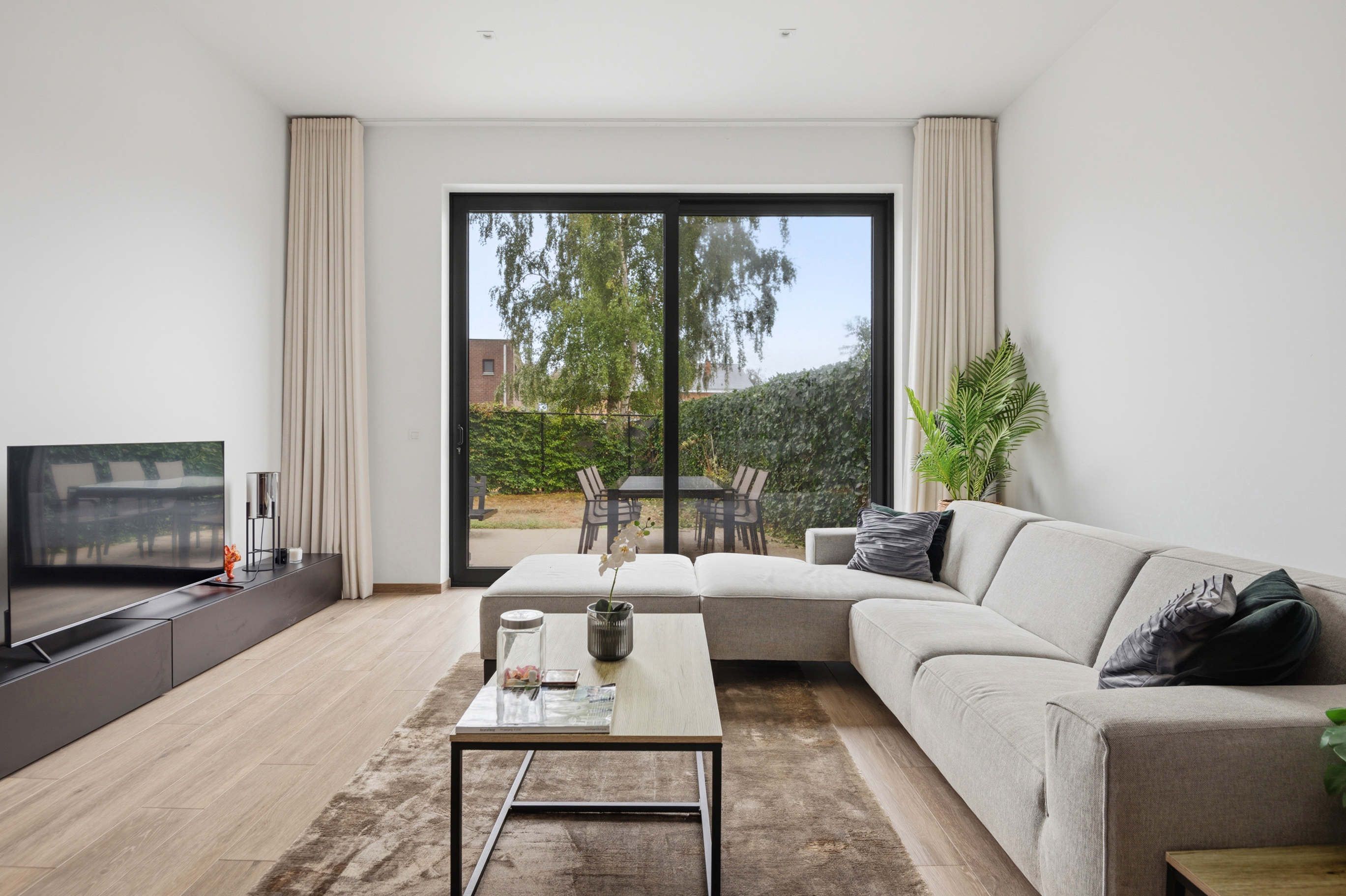 Modern living room with a light gray sectional sofa, coffee table, and large sliding doors opening to a patio and garden. [AI-generated text]