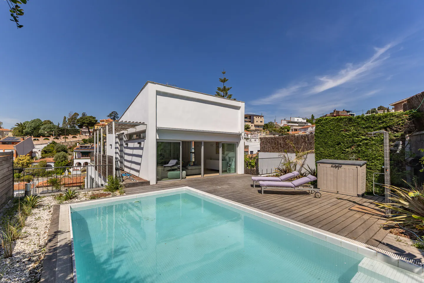 Modern white house with a pool and wooden deck. Two purple lounge chairs sit near a storage box and outdoor shower. Blue sky above.